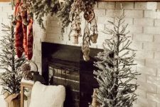 a rustic Christmas mantel wih a snowy evergreen garland with lights, stockings and antlers and mini Christmas trees next to the fireplace