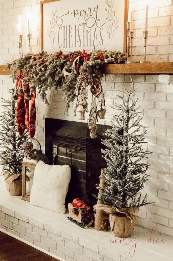 a rustic Christmas mantel wih a snowy evergreen garland with lights, stockings and antlers and mini Christmas trees next to the fireplace
