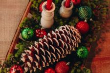 a woven bowl with evergreens, red and green ornaments, a large snowy pinecone and tall and thin red candles