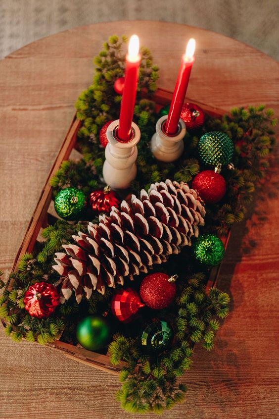 a woven bowl with evergreens, red and green ornaments, a large snowy pinecone and tall and thin red candles