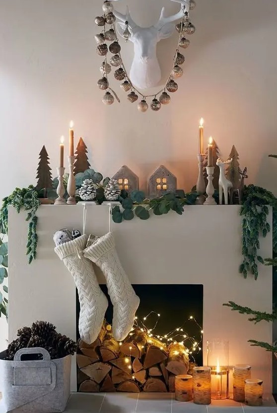 an elegant Scandinavian Christmas mantel with wooden Christmas trees, a greenery garland, candles in wooden candleholders, mini houses and snowy pinecones