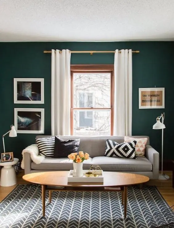 a catchy small living room with dark green walls, a grey sofa with printed pillows, a printed rug, an oval table and some artwork