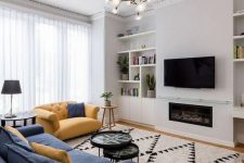 a modern and bright living room with a TV and a fireplace, built-in shelves, a blue and a yellow sofa, coffee tables and a chandelier
