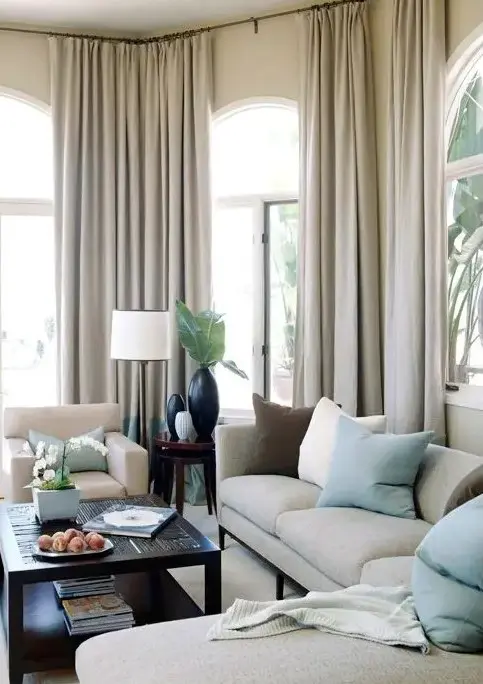 A refined neutral living room with arched windows treated with neutral curtains, a sectional with neutral and mint colored pillows and a dark coffee table for a contrast