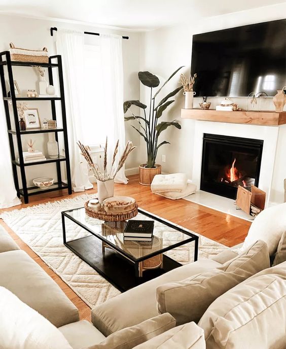 a small and inviting living room with a fireplace, a neutral sectional, a shelving unit, a coffee table, potted plants and pillows