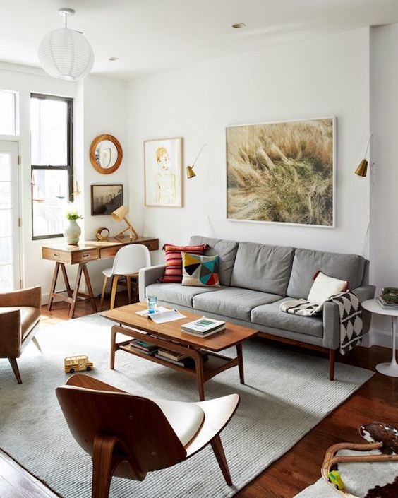 A small mid century modern living room with a grey sofa, a tiered coffee table, a leather chair and a neutral one, a desk and a white chair, some art