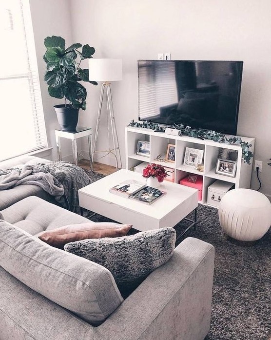 a small yet cozy living room with a small grey sofa, a TV unit, a white table and some lamps plus a potted plant