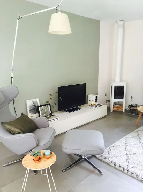 a stylish living room with a light green accent wall, a TV unit and a TV, a white hearth, a grey chair with an ottoman, some decor and a lamp