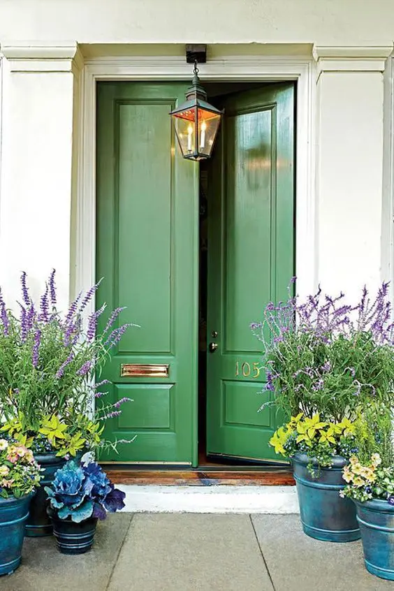 a beautiful porch with green doors, gold numbers, a hanging lantern, potted blooms and greenery is cool