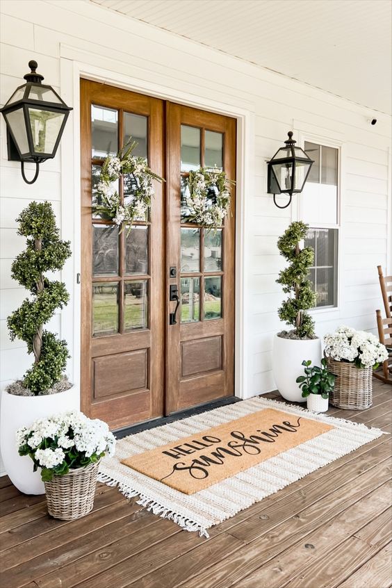 a beautiful spring farmhouse porch with layered rugs, potted blooms and greenery, greenery and white flower wreaths