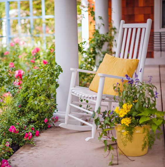 a bold porch with a white rocker and a yellow pillow, potted bright blooms and greenery is amazing