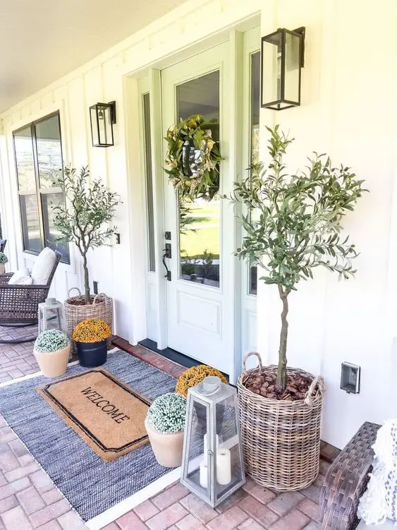a cool spring porch styled with trees in baskets, potted blooms, layered rugs and lanterns and a greenery wreath