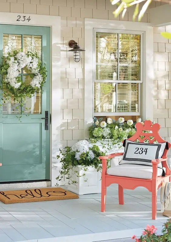 a lively spring porch with lush white blooms and greenery in planters, a coral chair with a pillow, a white bloom and greenery wreath