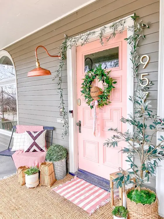 a lovely spring cottage porch with a pink door, a rose gold lamp, greenery in pots and a green wreath, layered rugs and a bench with pillows