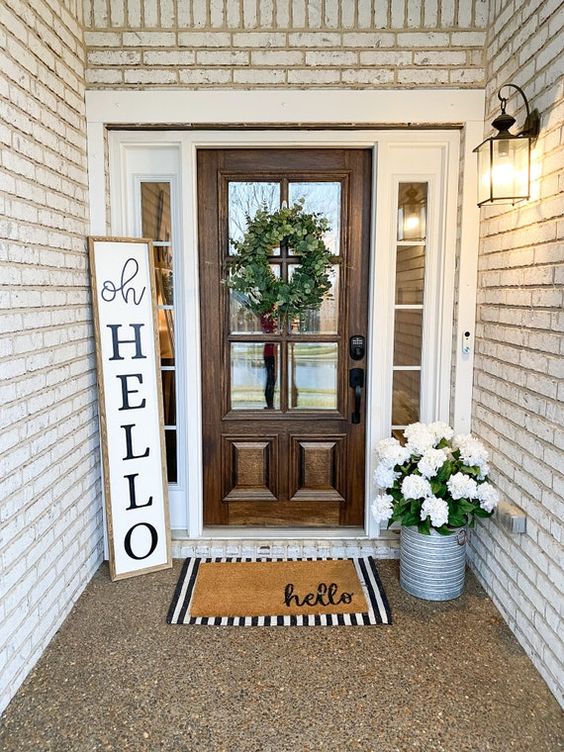 a neutral spring porch with potted white blooms, a sign, layered rugs and a greenery wreath is a lovely space