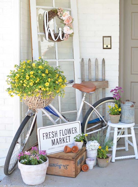 a spring cottage porch with a vintage door and a wreath, a bike with a basket of blooms, some potted plants and a piece of fence