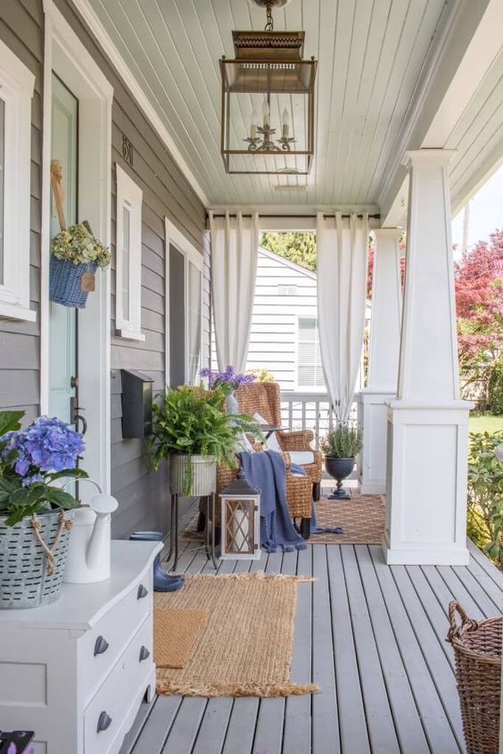 a welcoming spring porch with rugs, a woven chair with a footrest, potted plants and blooms plus some baskets