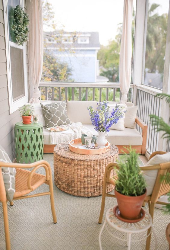 a welcoming spring porch with stained and rattan furniture, a woven pouf as a table, potted greenery and blooms