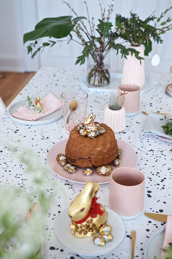 a cute modern Easter tablescape with a speckled tablecloth, pink and white porcelain, some greenery, vases and gold cutlery