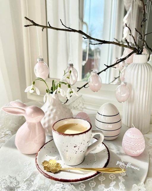 a modern Easter place setting with a neutral cup and saucer, a pink bunny, some eggs, branches with egg ornaments and some snowdrops in a vase