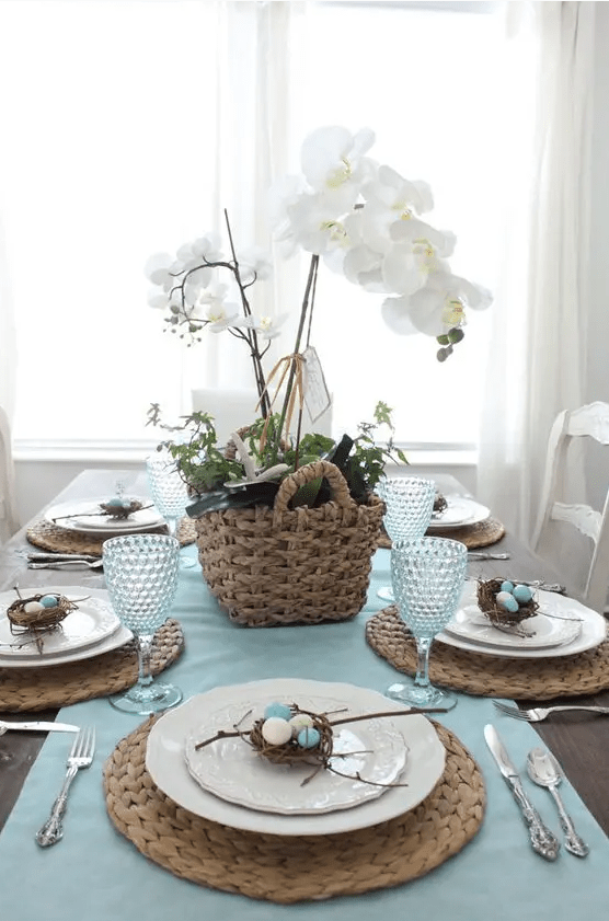 a rustic Easter tablescape with a blue runner, woven placemats, a basket with greenery and white orchids, mini nests with eggs