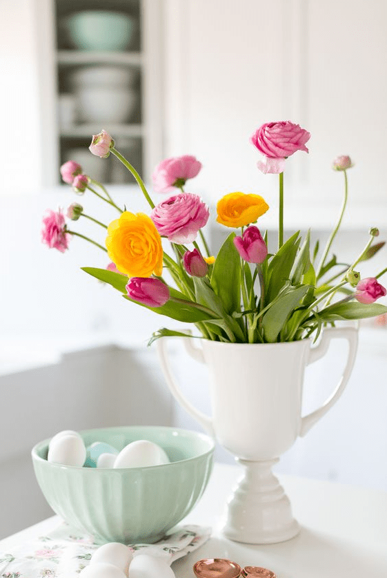 a spring centerpiece of a white cup with bold pink and yellow ranunculus and some eggs in a bowl