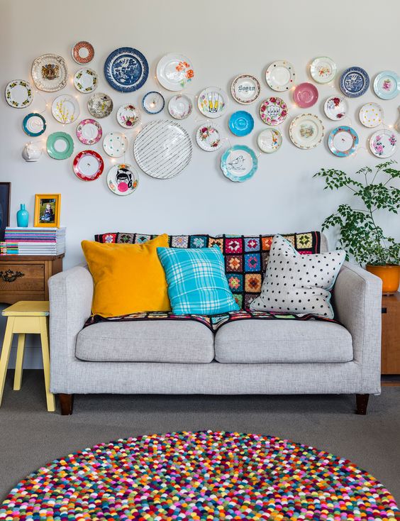 a colorful living room with a neutral sofa and bright pillows, a bold plate wall, a colorful pompom rug and some potted plants and artwork