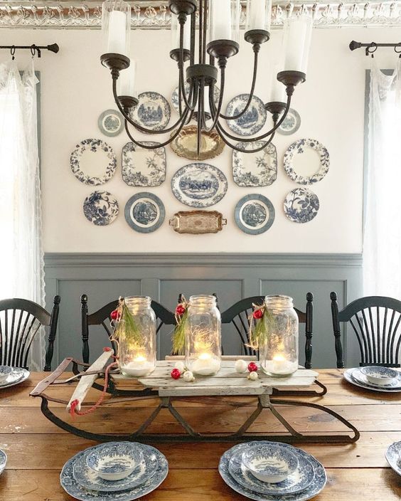a vintage farmhouse dining room with paneled walls, a stained table, black chairs, a vintage plate wall and a chic chandelier