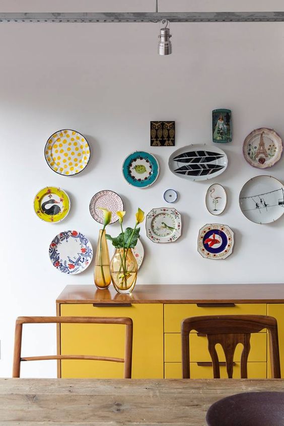 an eclectic dining room with a stained table and stained chairs, a yellow credenza and a quirky and colorful plate wall for decorating the space