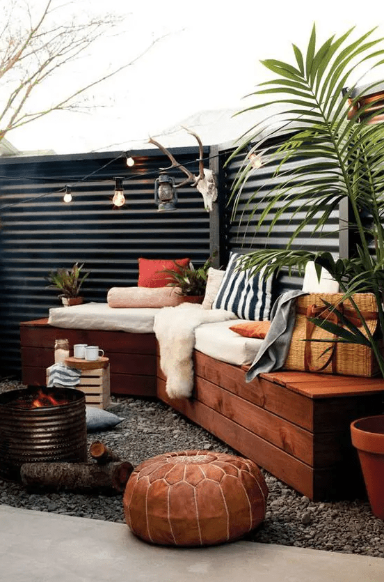 a black corrugated metal fence, a stained corner sofa, some pillows, lights, potted plants, a leather pouf and a fire pit
