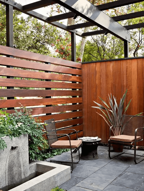A mid century modern patio with a wooden stained fence, modern chairs and a side table, potted plants and a modern fountain