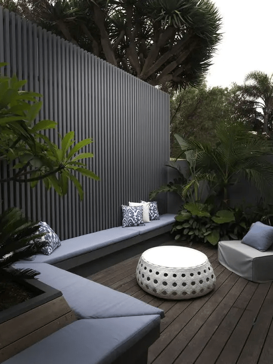 a modern deck with a built-in bench with cushions and pillows, greenery and trees, a modern chair and a coffee table and a grey timber screen