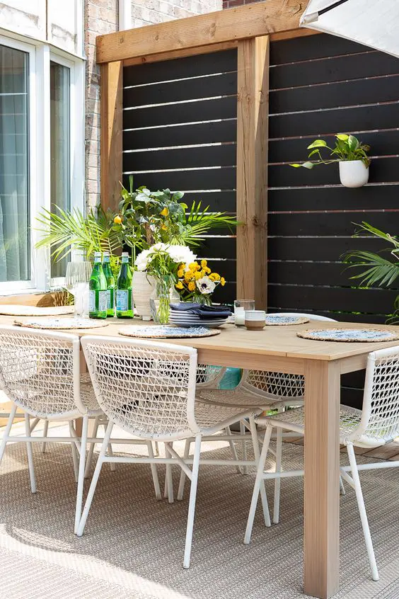 a modern terrace with a black and stained privacy screen, a stained table, white wire chairs, greenery and blooms is cool