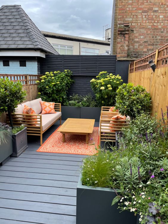 a modern terrace with a black fence and some privacy screens, raised beds and planters with greenery and blooms and stained furniture