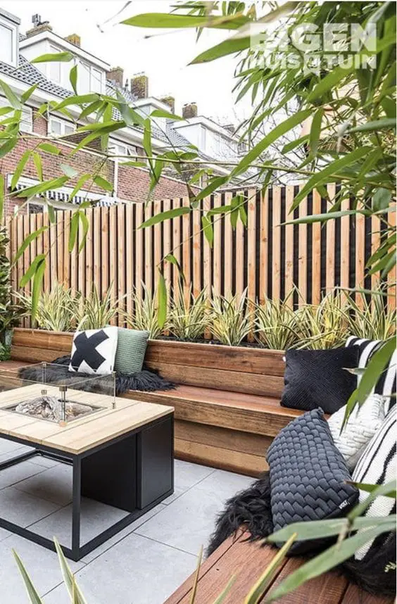 A modern terrace with a stained bench and pillows, a fire pit table, a built in planter with greenery and a privacy fence