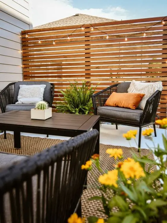 a modern terrace with a stained screen and lights, modern outdoor furniture, some greenery and blooms plus pillows