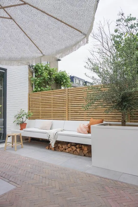 a neutral terrace with a stained screen, a white sofa and firewood, a planter with a tree, a stool and pillows
