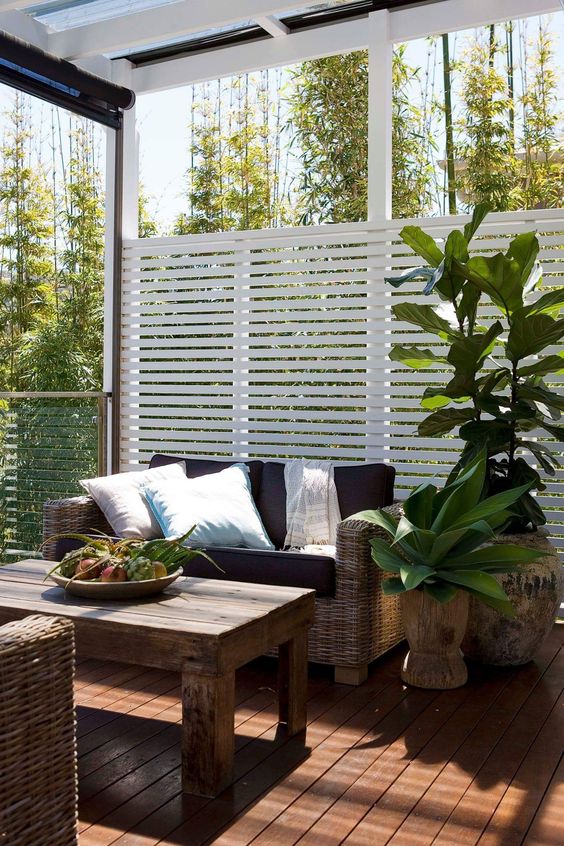 a small terrace with a privacy screen, wicker furniture and a wooden table, some woodne planters with large plants