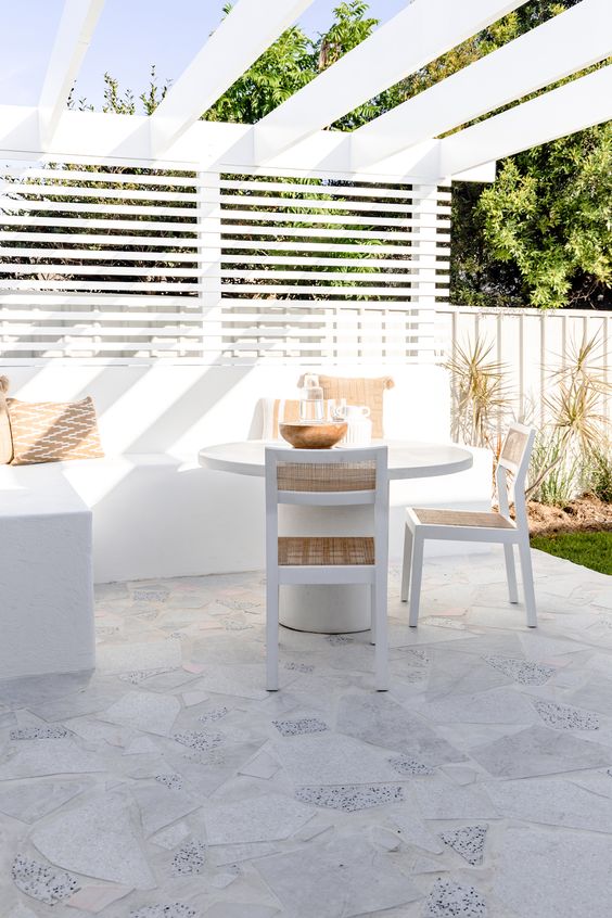 a white terrace with privacy screens, a white corner sofa, a table and chairs, some boho pillows and grasses around