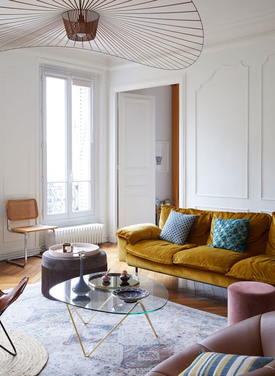 a Parisian living room with a mustard velvet sofa and pillows, a coffee table, a pink pouf, a cane chair and a leather chair plus a large chandelier