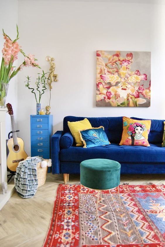 a bright living room with a navy sofa, a green velvet pouf, a printed rug, a basket with blankets, a blue file cabinet, some blooms and a bold artwork