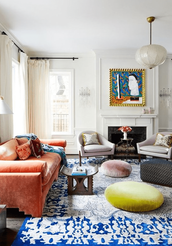 a colorful eclectic living room with a fireplace, lilac chairs, an orange velvet sofa, Moroccan furniture, a rug and poufs is bold
