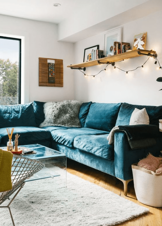 a cozy mid-century modern living room with a navy velvet sectional, an acrylic coffee table, a metal chair, a shelf with lights and some pillows