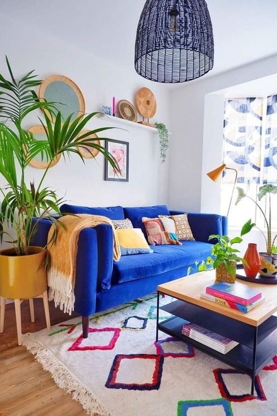 a dopamine decor living room with a blue velvet sofa and pillows, a coffee table, a colorful rug, potted plants, a ledge with decor and a gallery wall