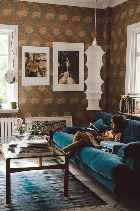 a gorgeous eclectic living room with bold floral wallpaper, a modern turquoise sofa, a glass table and a pretty gallery wall