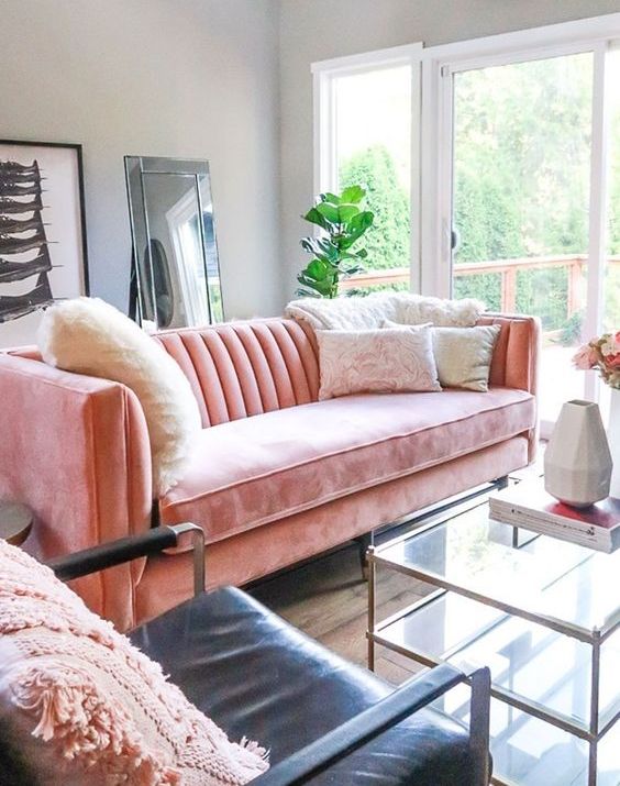 A light filled living room with a coral velvet sofa, a tiered coffee table, a black leather chir, some decor and a floor mirror
