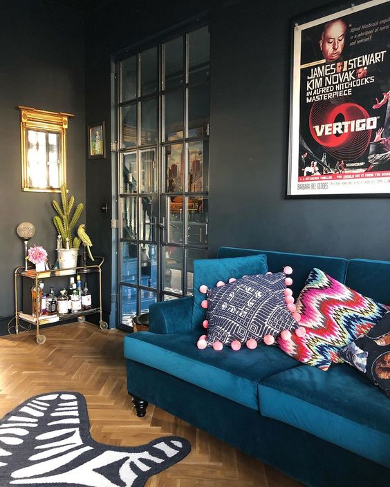 a moody living room with graphite grey walls, a navy velvet sofa with printed pillows, some art, a rug and a bar cart in the corner