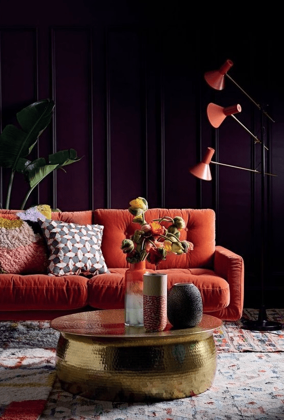 a moody living room with purple paneled walls, an orange sofa, orange floor lamps, a gold hammered table and statement plants