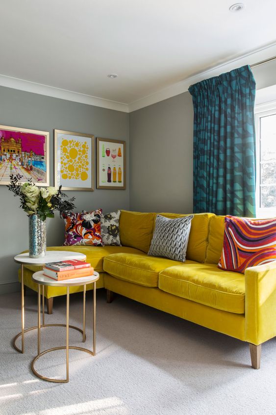 a neutral living room with grey walls, a sunny yellow velvet sofa, colorful pillows and curtains, a duo of coffee tables, a bold gallery wall