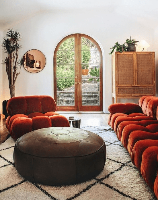 a refined living room in neutrals, with a burnt orange sofa and a chair, a round leather ottoman, a rattan sideboard